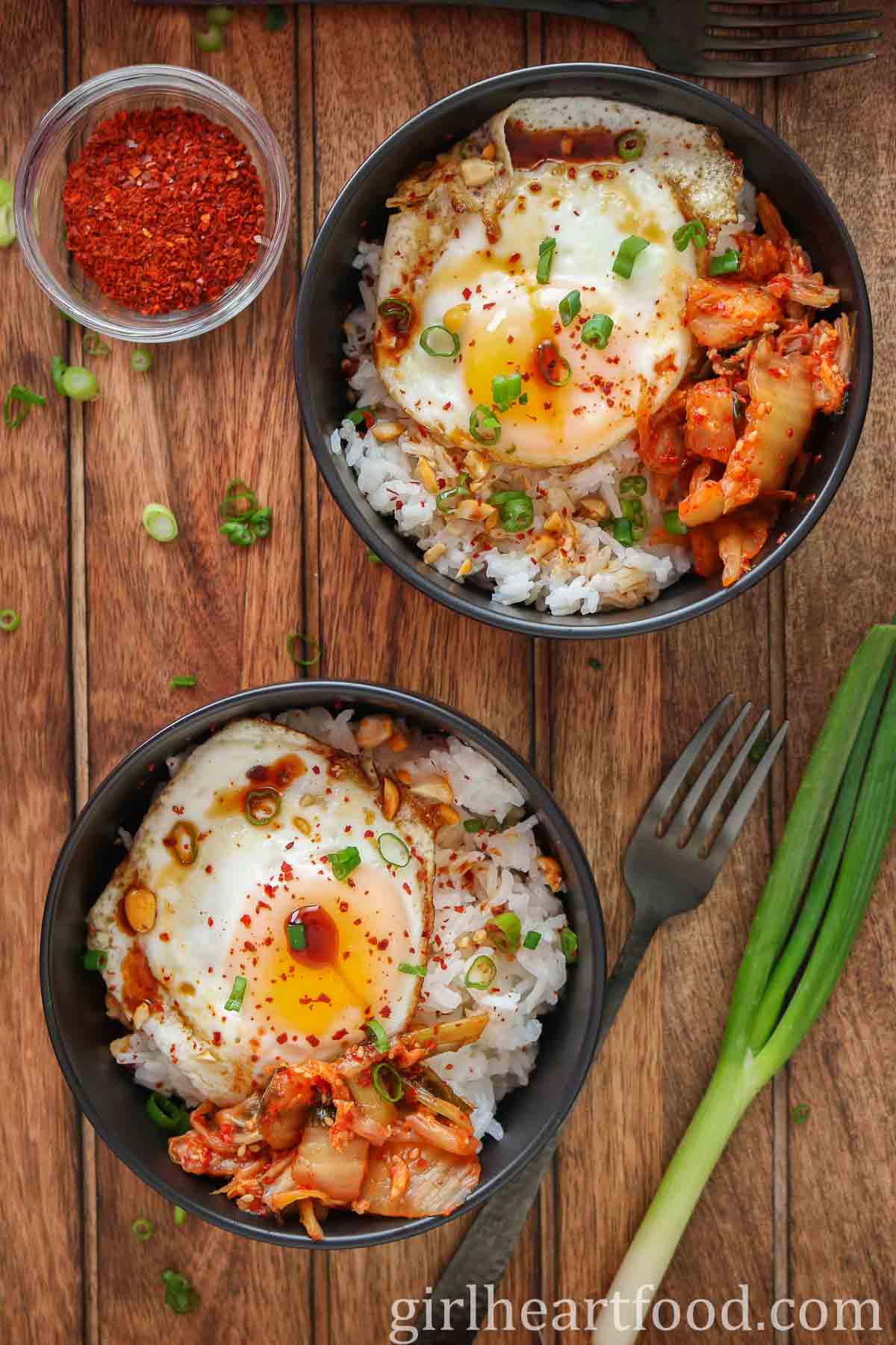 Two fried egg and rice bowls alongside green onion and a small dish of red chili pepper flakes.