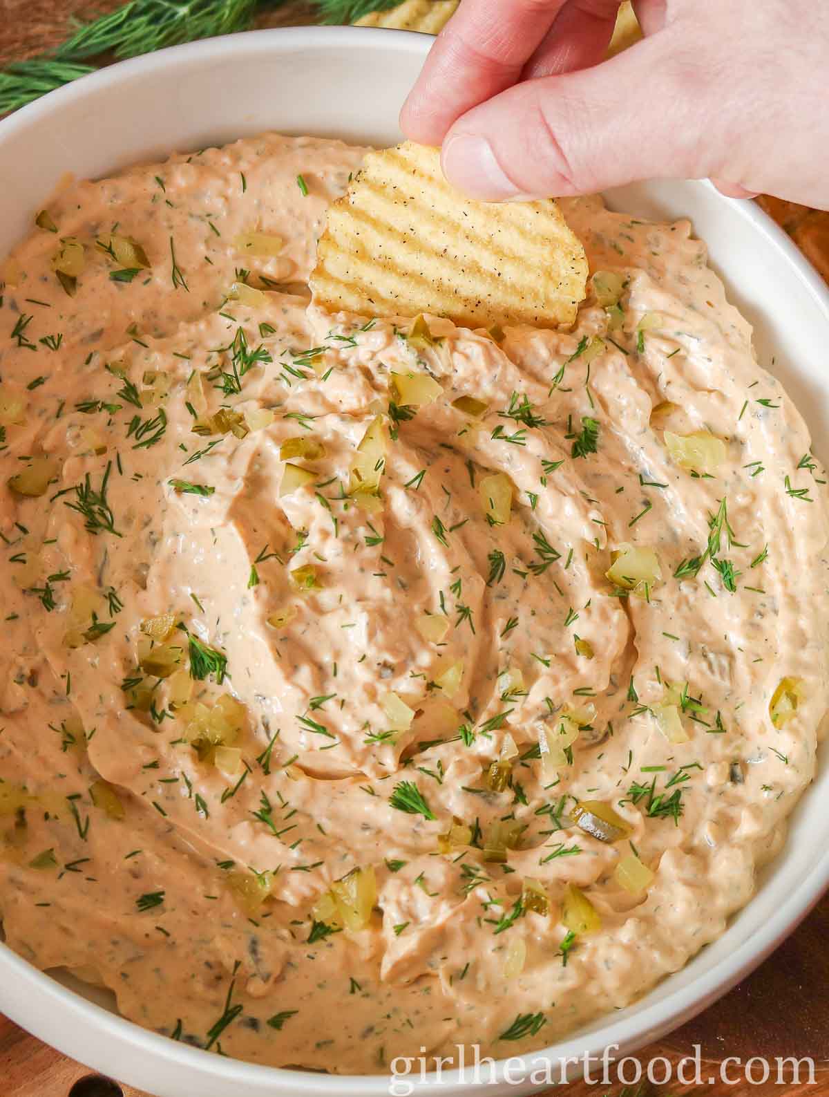 Hand dipping a potato chip into a bowl of dill pickle dip.