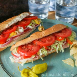Two Italian cold cut submarine sandwiches with two glasses of water behind them.