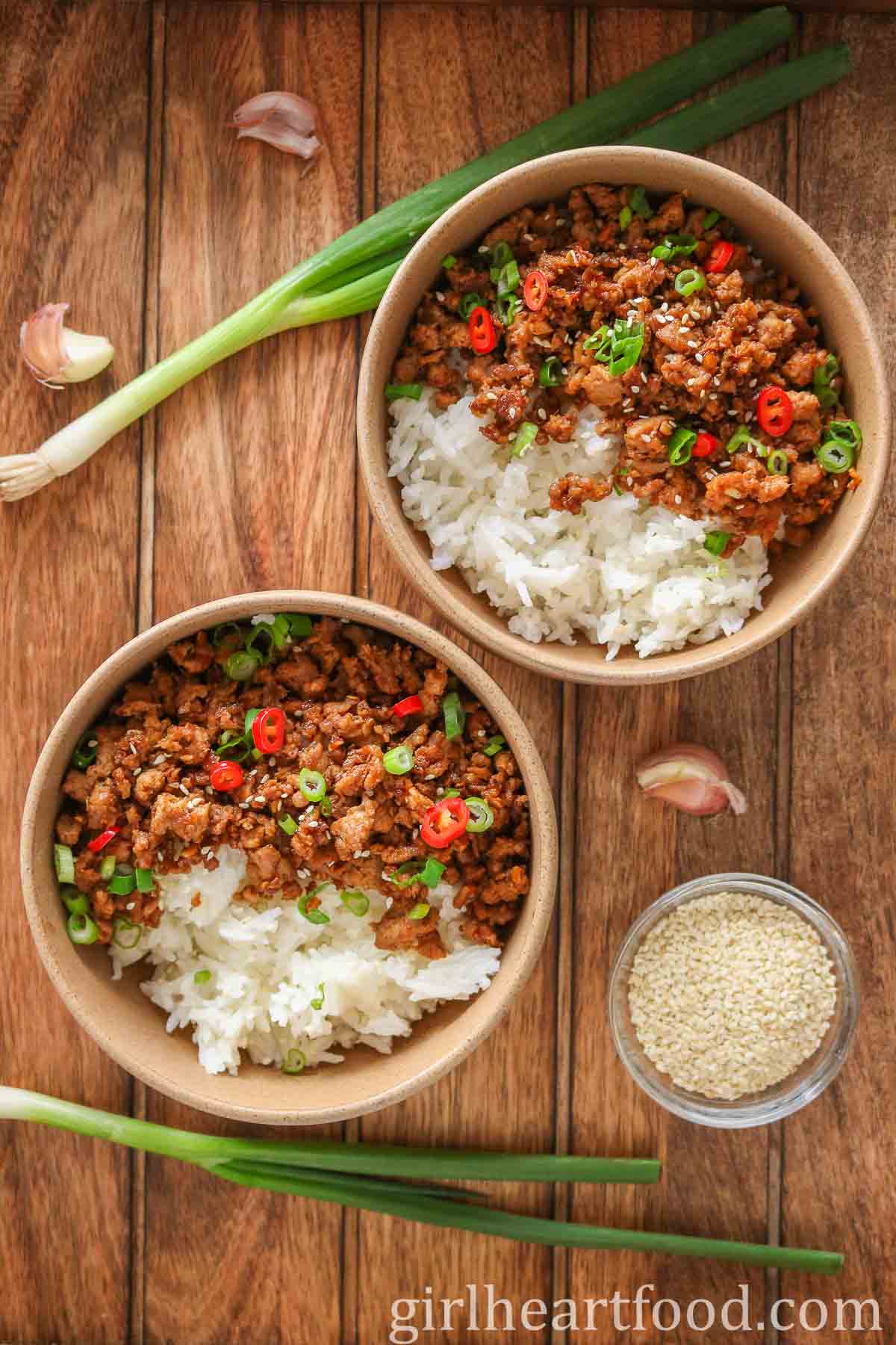 Two ground turkey and rice bowls next to green onion, garlic cloves and sesame seeds.