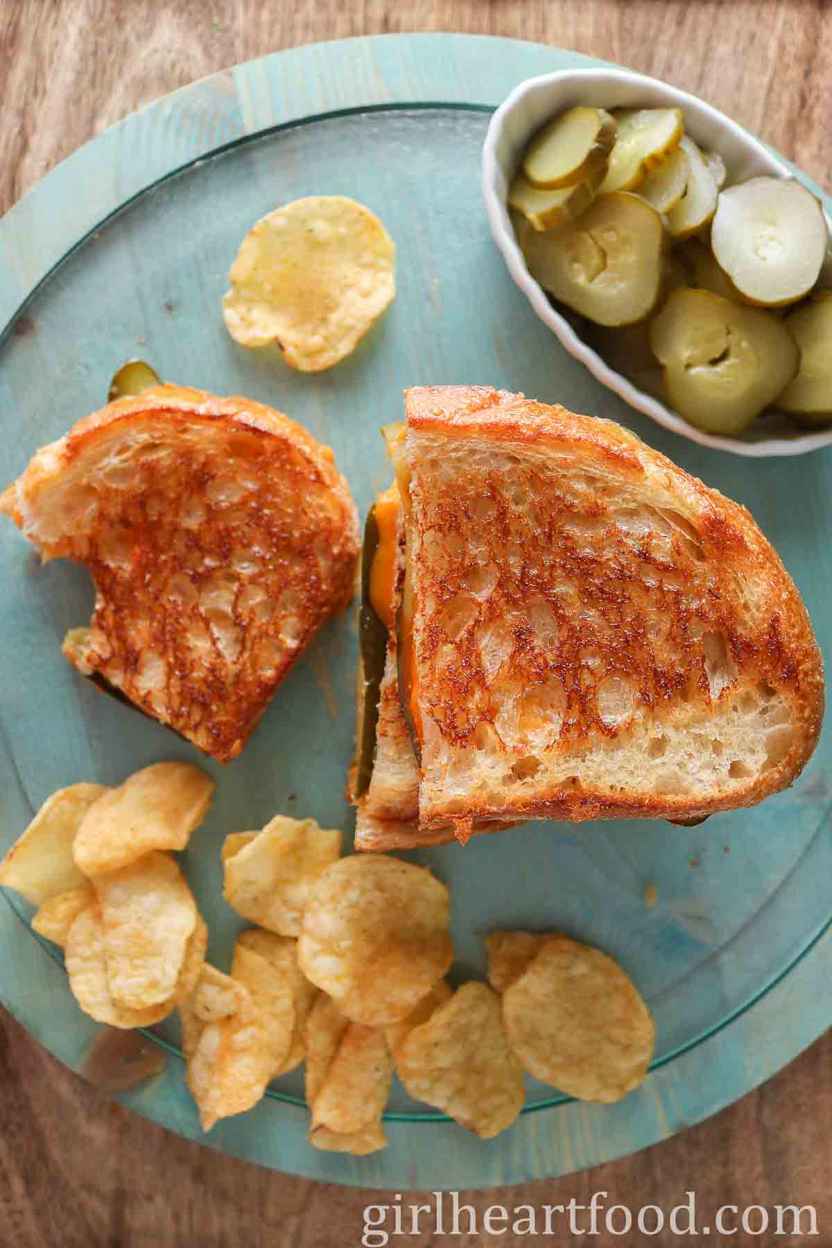 Grilled cheese sandwiches, potato chips and dish of sliced pickle on a round blue board.