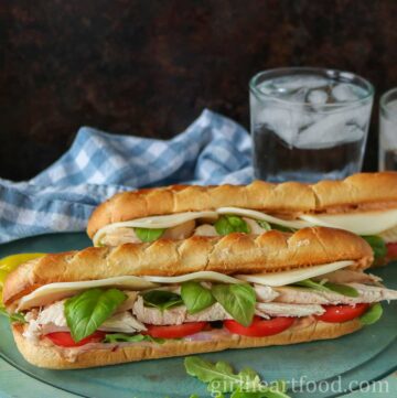 Two chicken baguette sandwiches in front of a tea towel and glass of water.