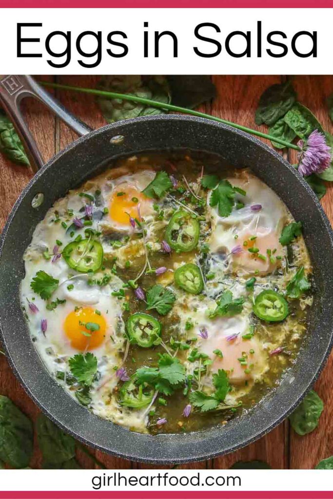 Pan of eggs in salsa verde garnished with toppings, and surrounded by spinach and chive flowers.
