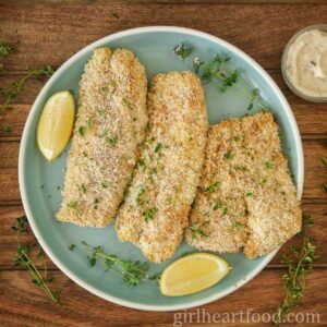 Three panko cod fillets with thyme and lemon wedges on a blue plate next to tartar sauce.