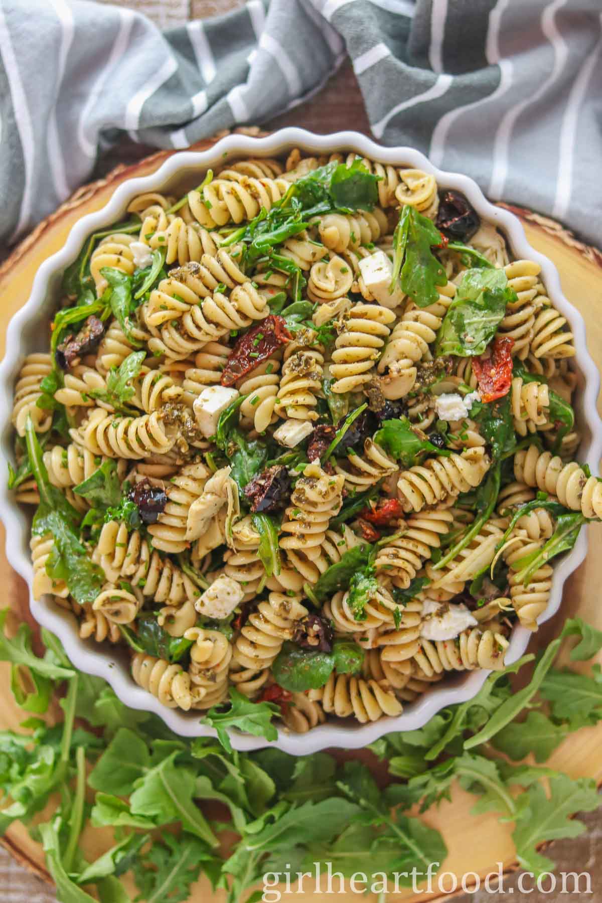 Bowl of Mediterranean pesto pasta salad next to a tea towel and arugula.