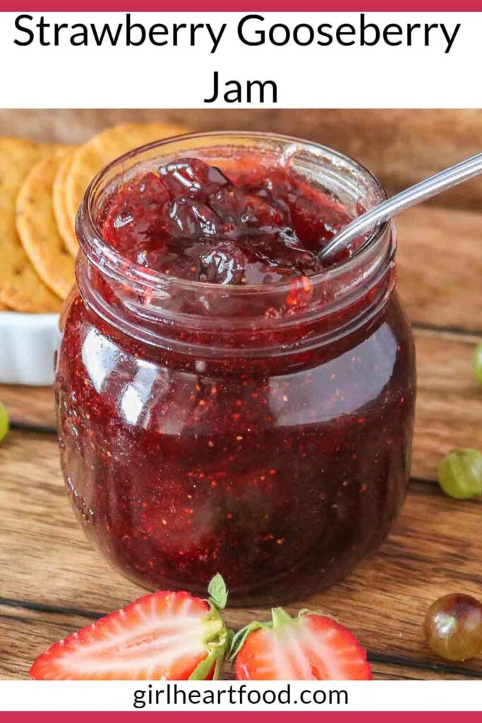 Jar of strawberry gooseberry jam with a spoon, next to a cut strawberry, gooseberries and crackers.