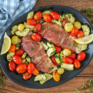 Prosciutto wrapped cod fillets, tomatoes, zucchini and lemon wedges on a serving platter.