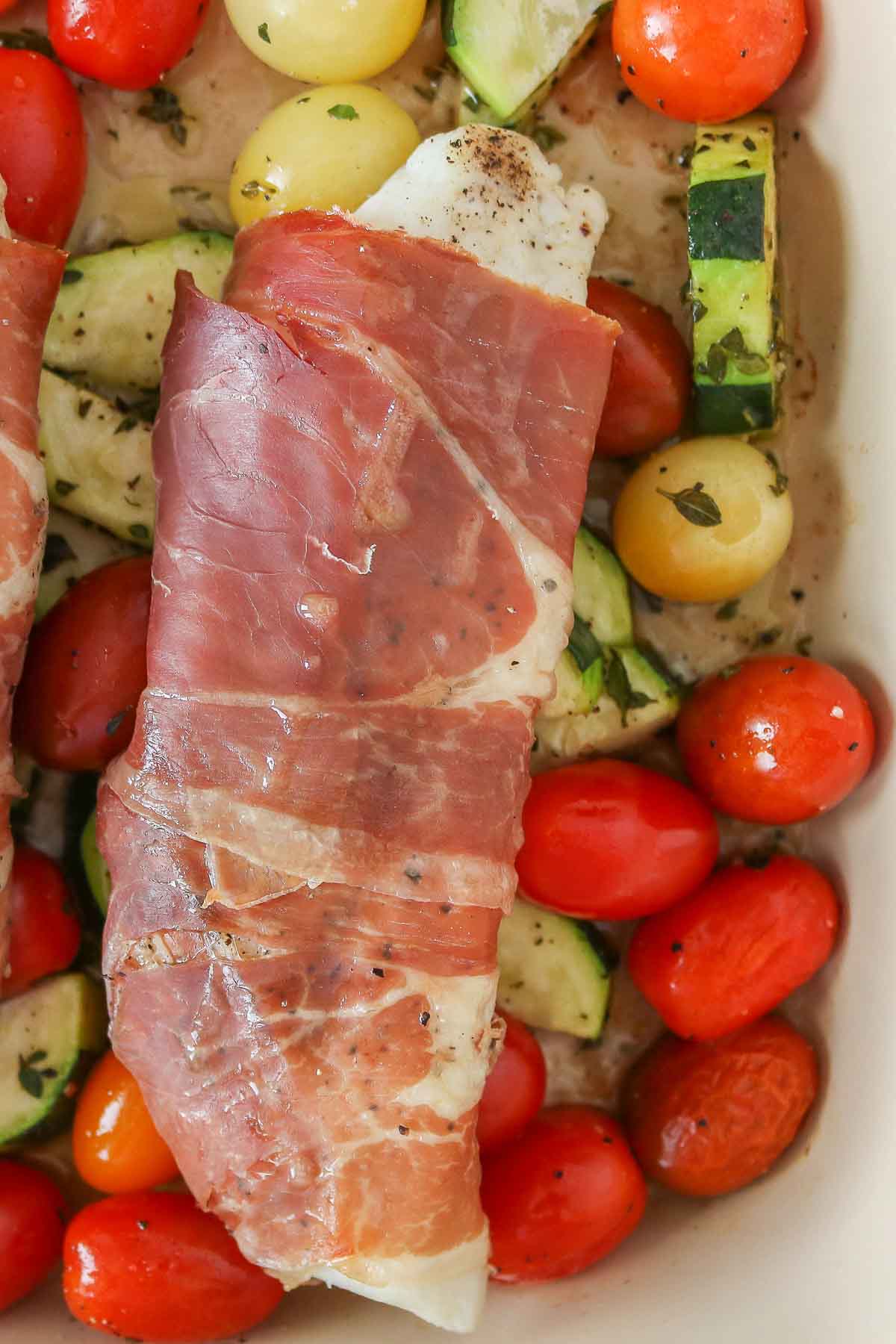 Close-up of a prosciutto wrapped cod fillet over tomatoes and zucchini.