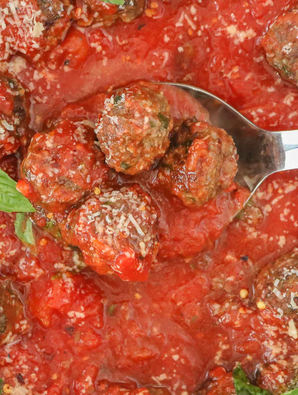 Serving spoon scooping up four baked meatballs with sauce from a pan.