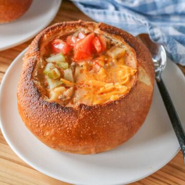 Cheeseburger soup in a bread bowl garnished with pickles, tomatoes and cheese.