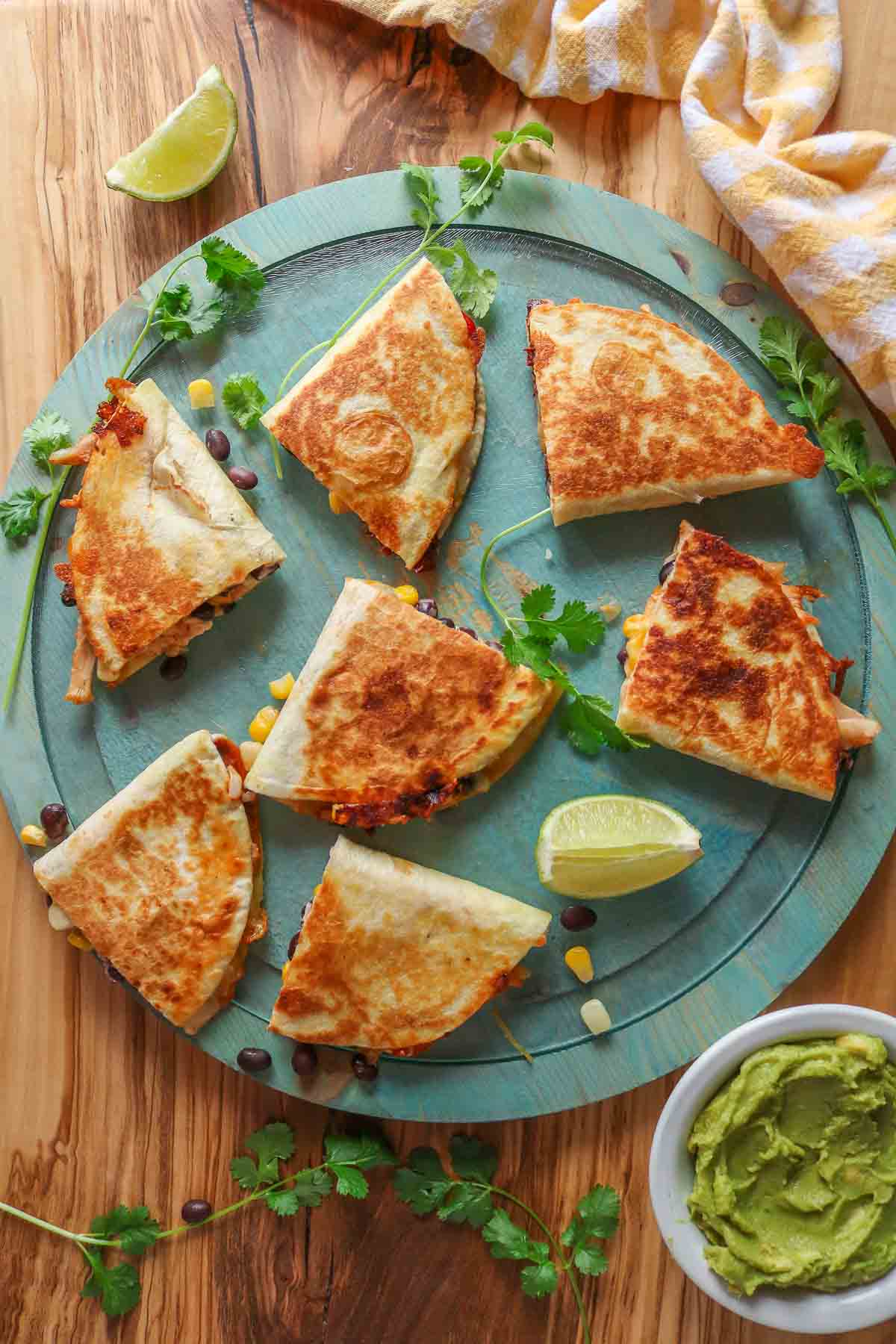 Quesadillas on a round blue platter next to lime wedges, cilantro and a dish of guacamole.