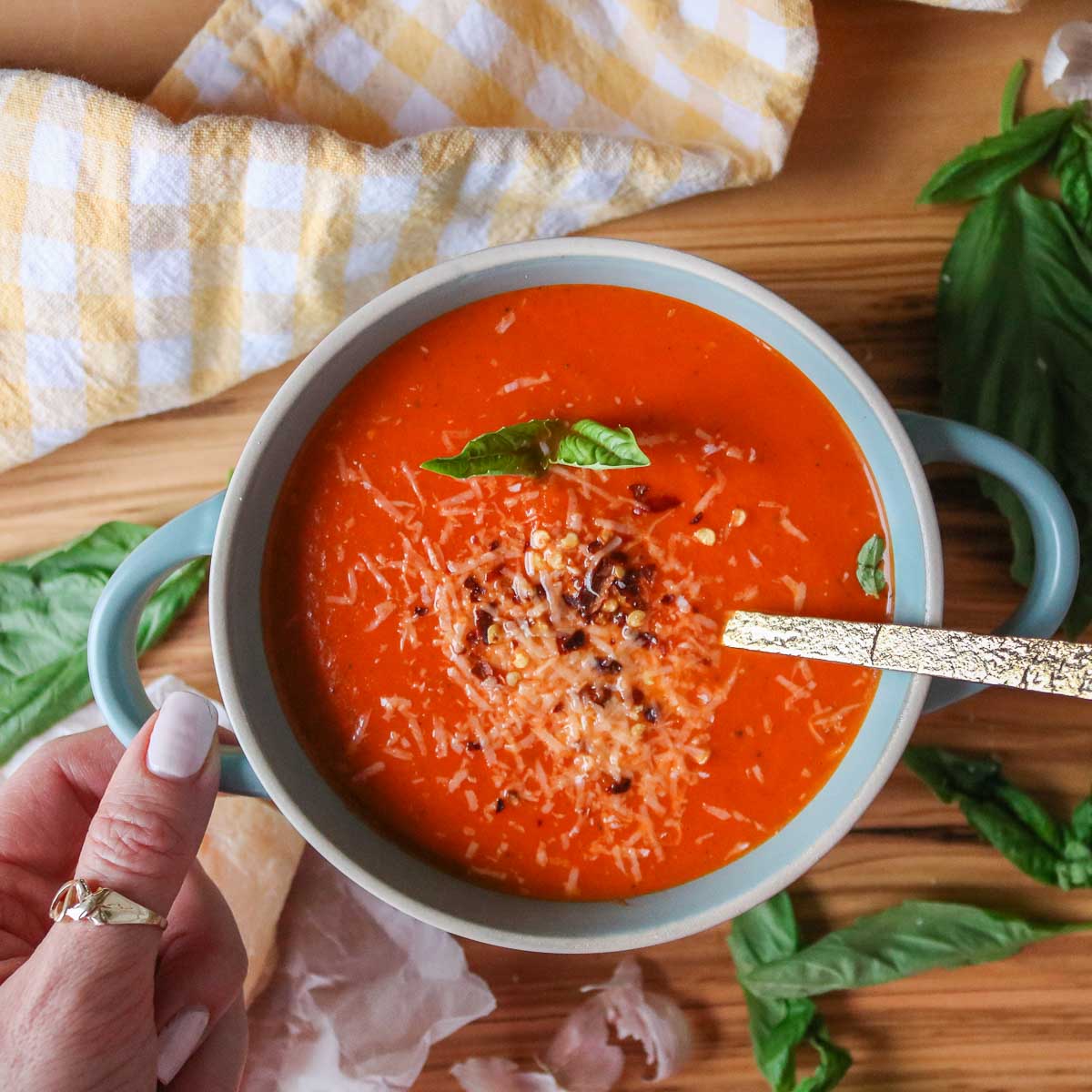 Hand holding a bowl of creamy tomato basil soup garnished with cheese, red pepper flakes and basil.
