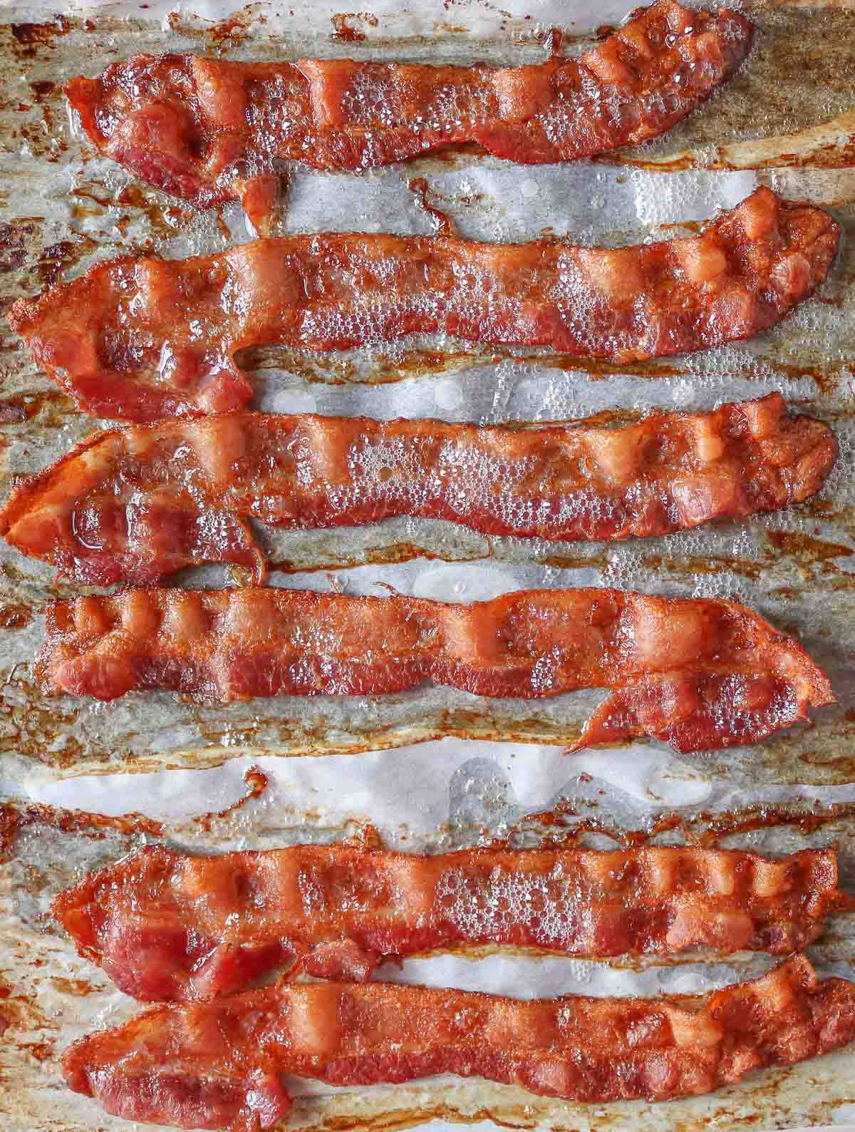 Six slices of crispy baked bacon on a parchment paper-lined sheet pan.