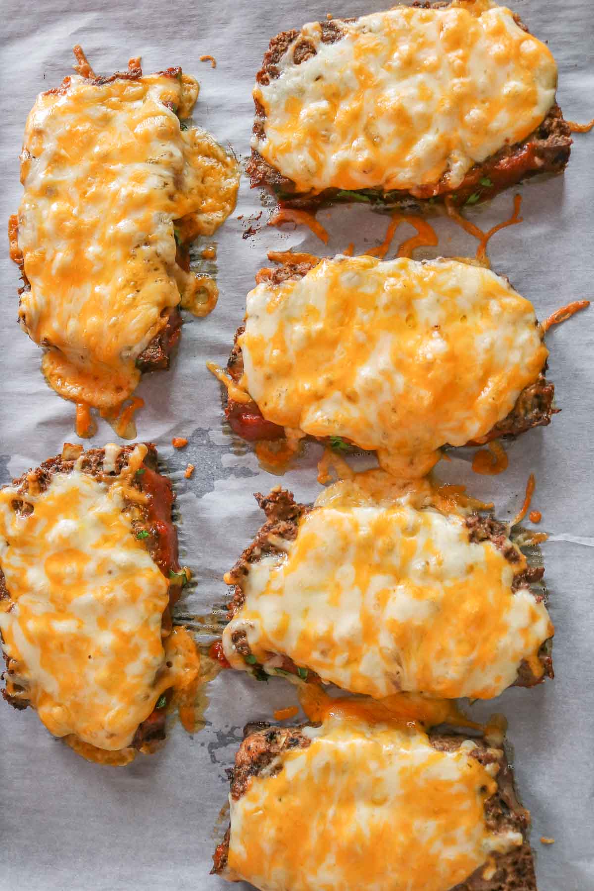 Six cheesy meatloaf slices on a parchment paper-lined sheet pan.