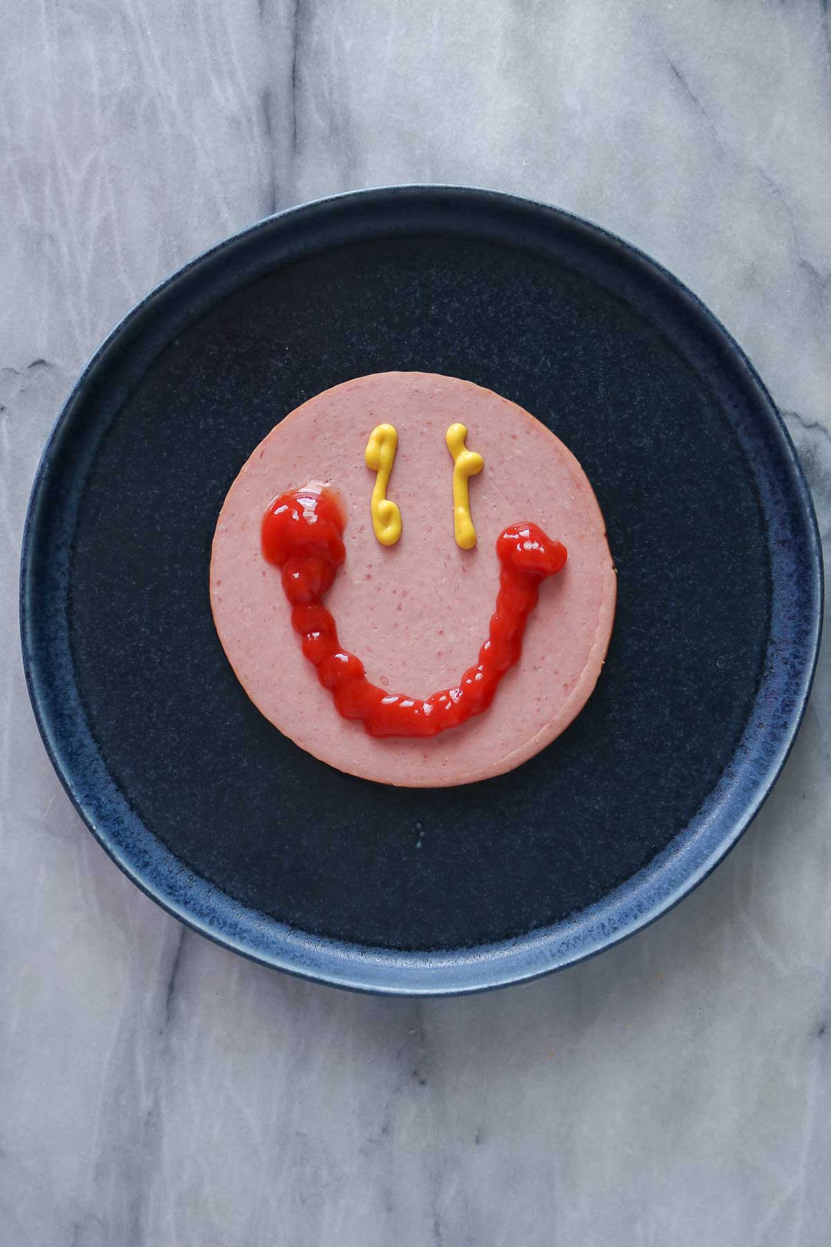 Slice of bologna on a blue plate with a smiling face drawn on it with mustard and ketchup.