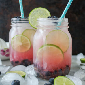 Two lime blueberries mocktails in glass jars, each garnished with blueberries and lime.