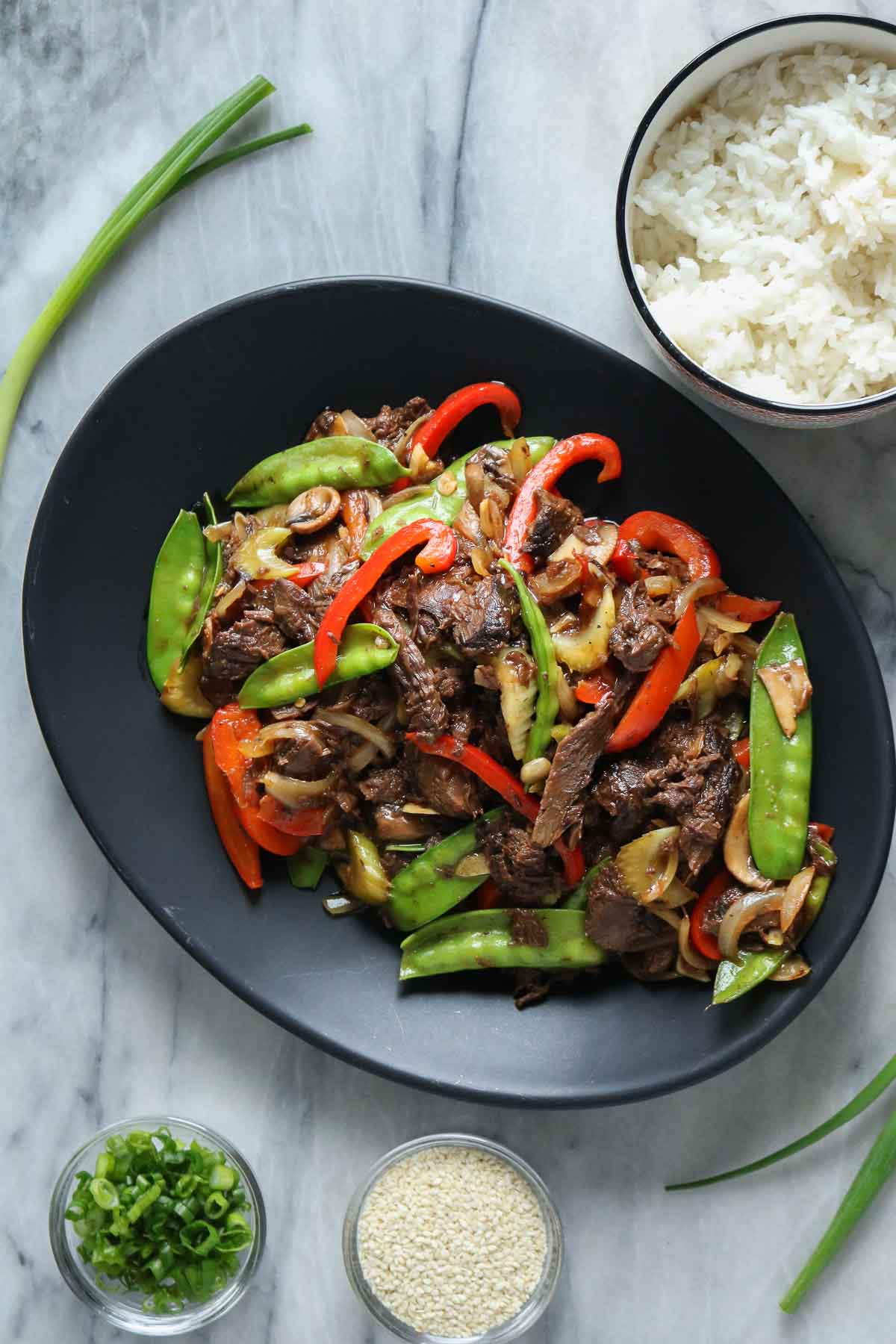 Moose stir-fry on a serving platter next to dishes of rice, green onion and sesame seeds.