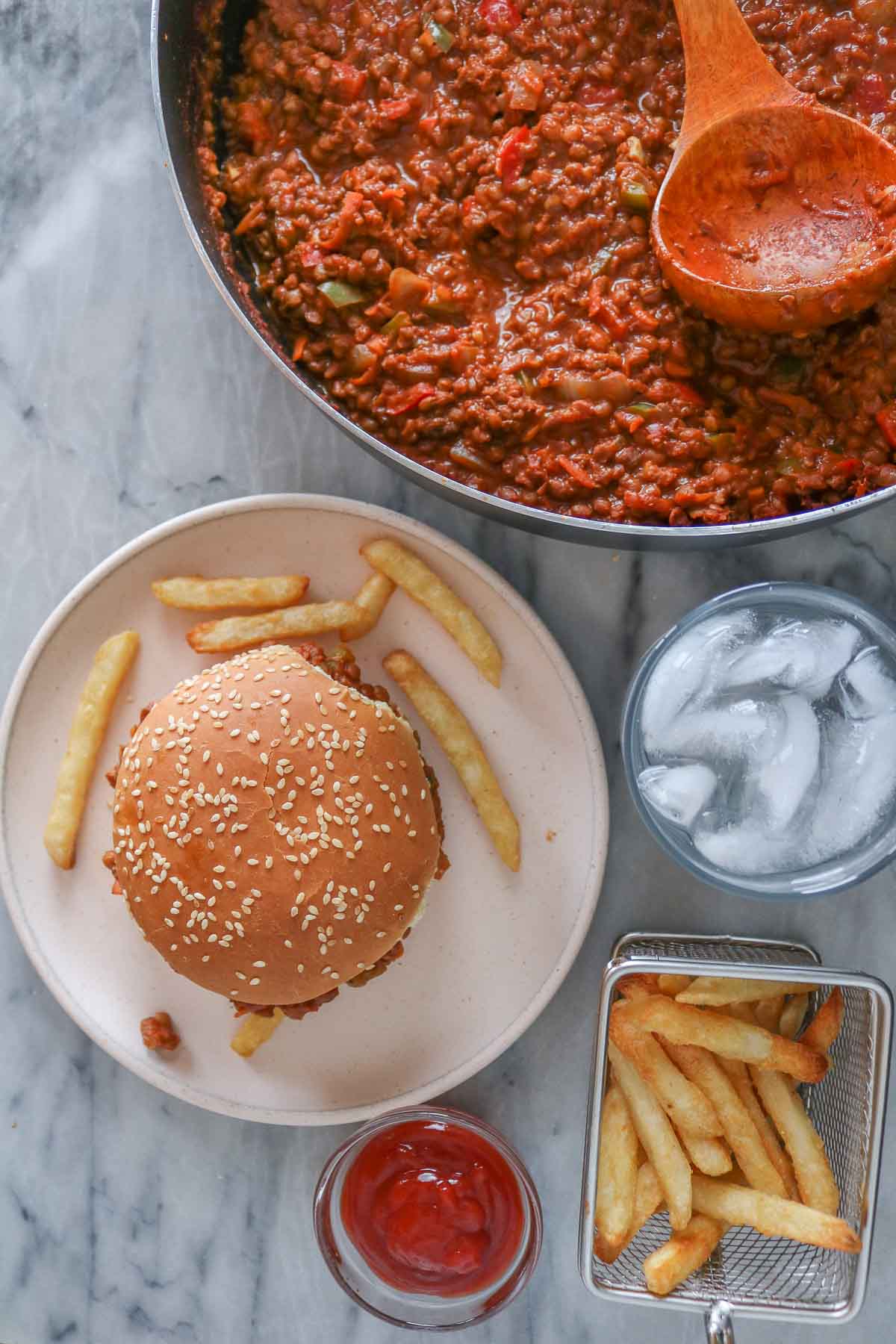 Overhead shot of a sloppy joe and fries next to a pan of the mixture, ketchup and glass of water.