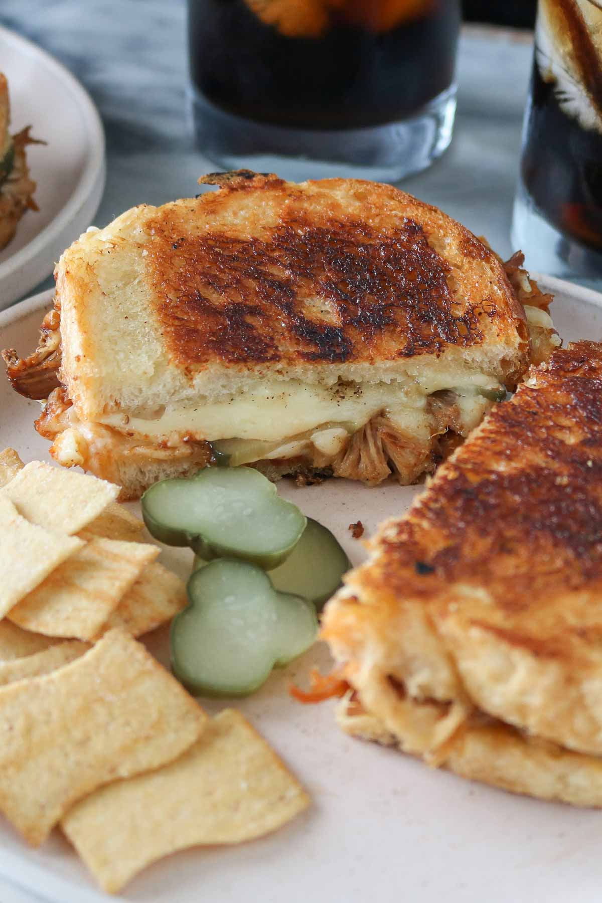 Two halves of a pulled pork grilled cheese sandwich on a plate with chips and pickle slices.