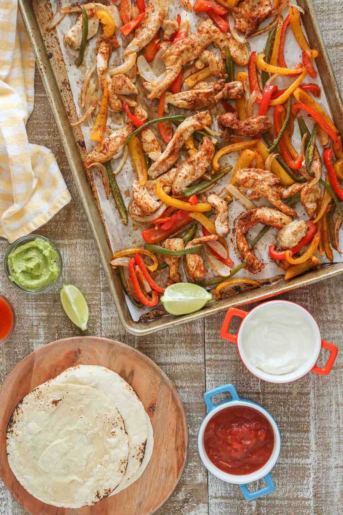 Cooked seasoned chicken, onion and peppers for fajitas on a sheet pan next to tortillas & toppings.