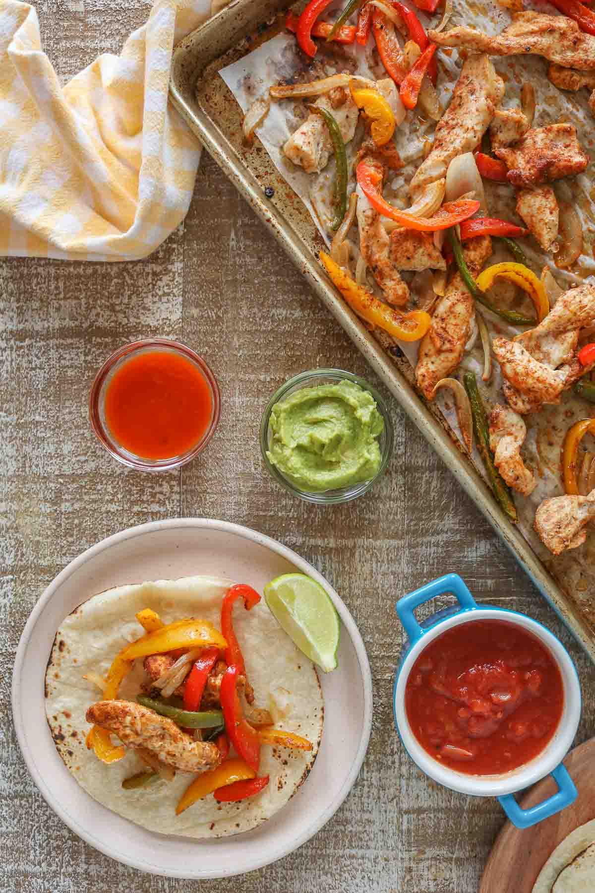 Chicken fajita on a plate next to toppings and a sheet pan with some of the chicken fajita mixture.