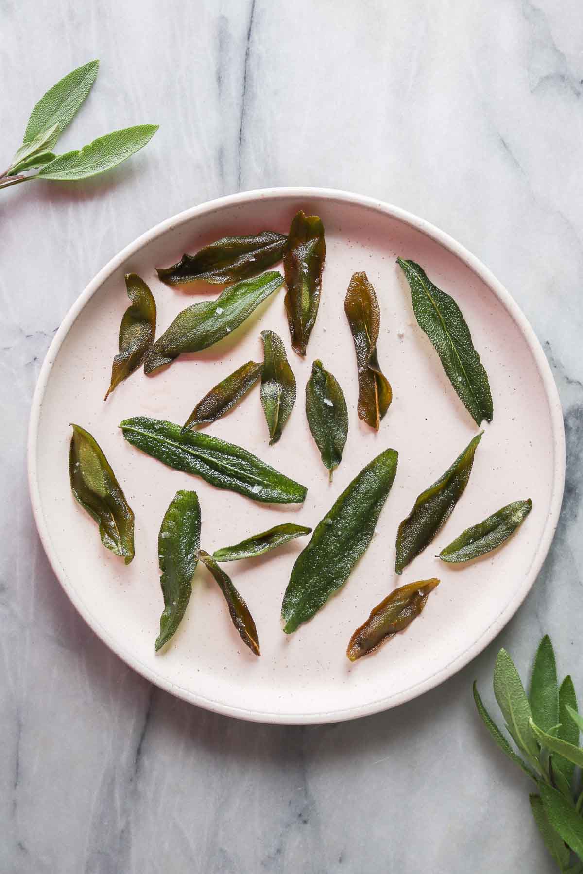 Crispy sage leaves on a plate surrounded by fresh sage leaves.