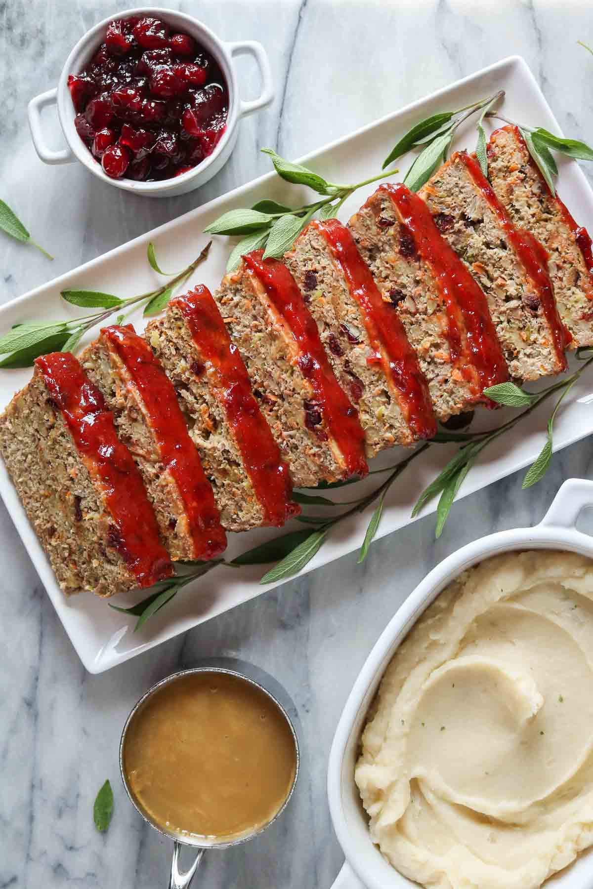 Sliced turkey meatloaf on a serving platter alongside mashed potatoes, gravy and cranberry sauce.