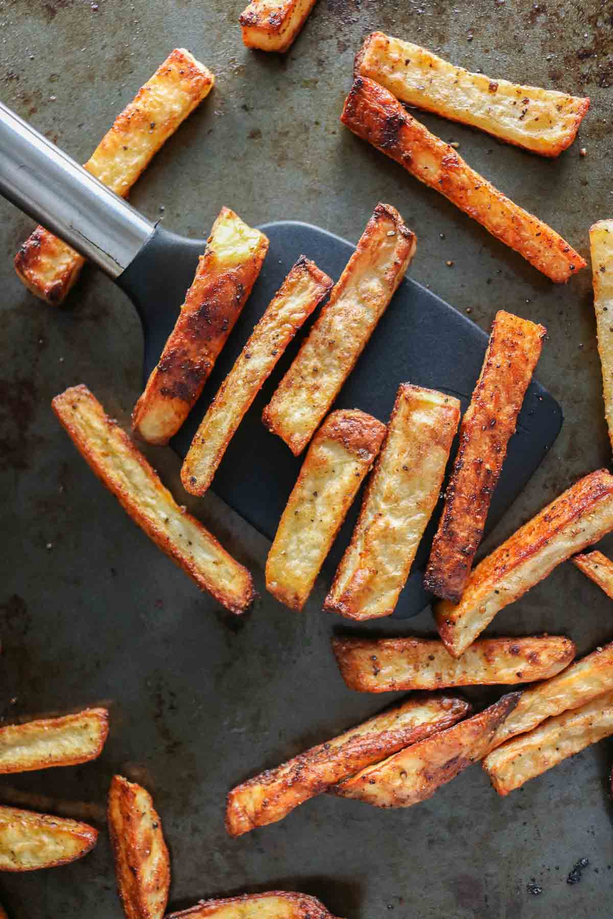 Spatula scooping up baked fries from a sheet pan.