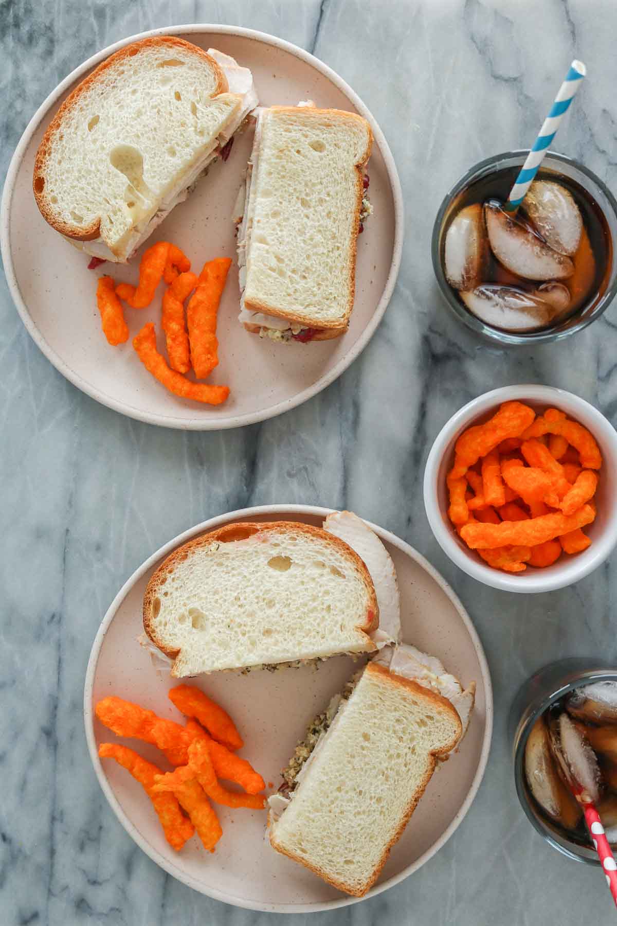 Two turkey and dressing sandwiches on plates with crunchy cheese puffs and glasses of soda.