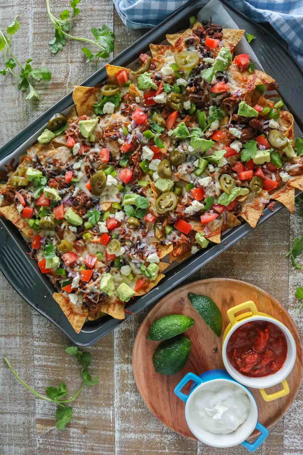 Pulled pork nachos on a sheet pan next to lime wedges and dishes of sour cream and salsa.
