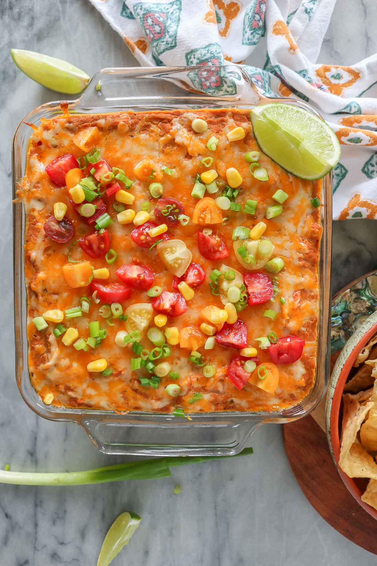 Cheesy refried bean dip in a baking dish with toppings alongside a bowl of tortilla chips.