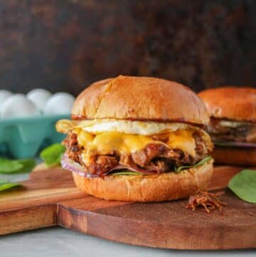 Pulled pork breakfast sandwiches on a serving board with a carton of eggs in the background.