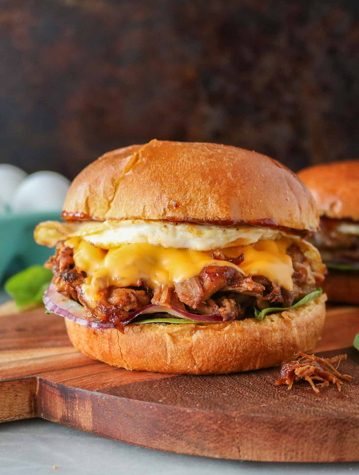 Pulled pork breakfast sandwiches on a serving board with a carton of eggs in the background.