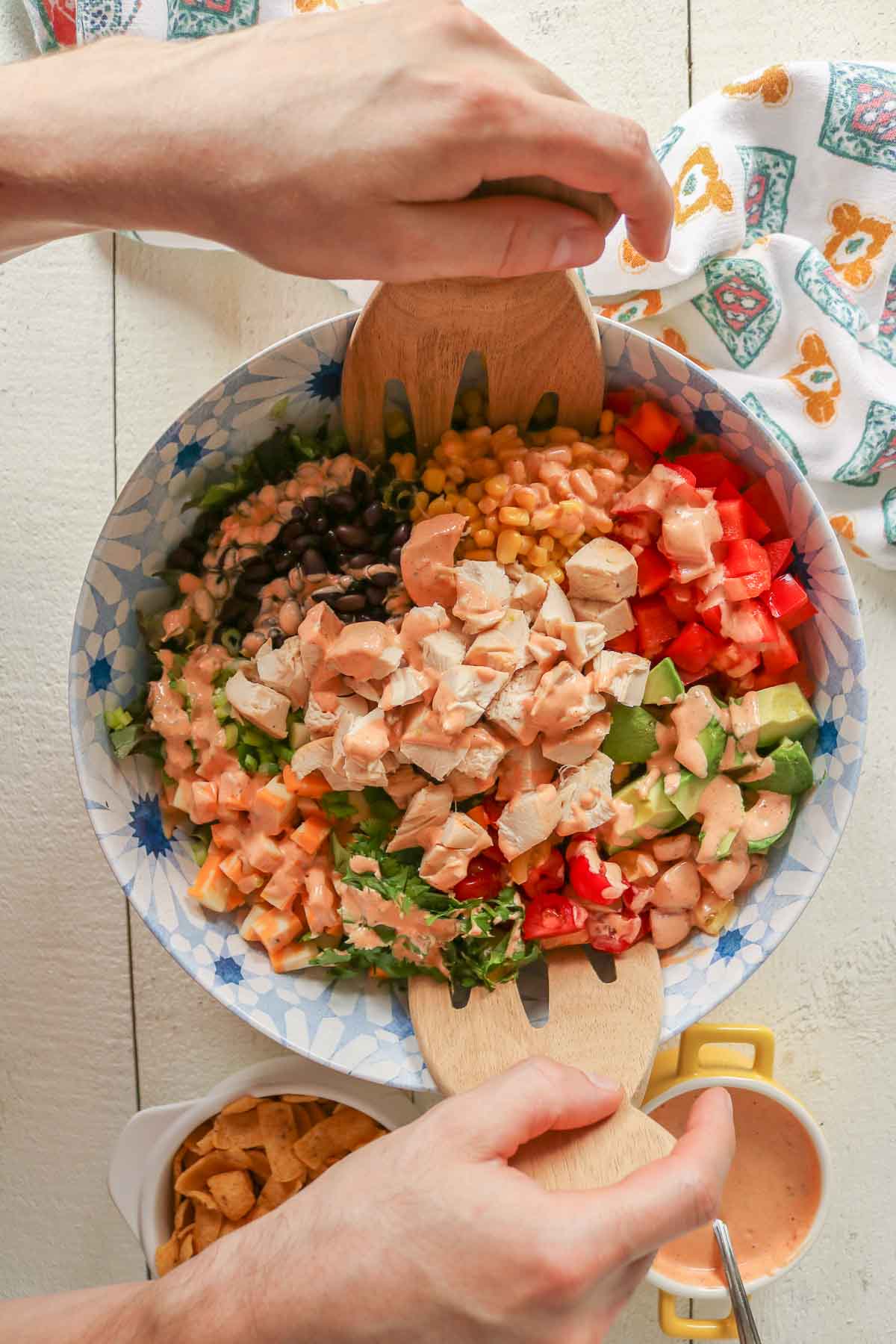 Two hands using salad tossers to mix salad ingredients and dressing together in a serving bowl.