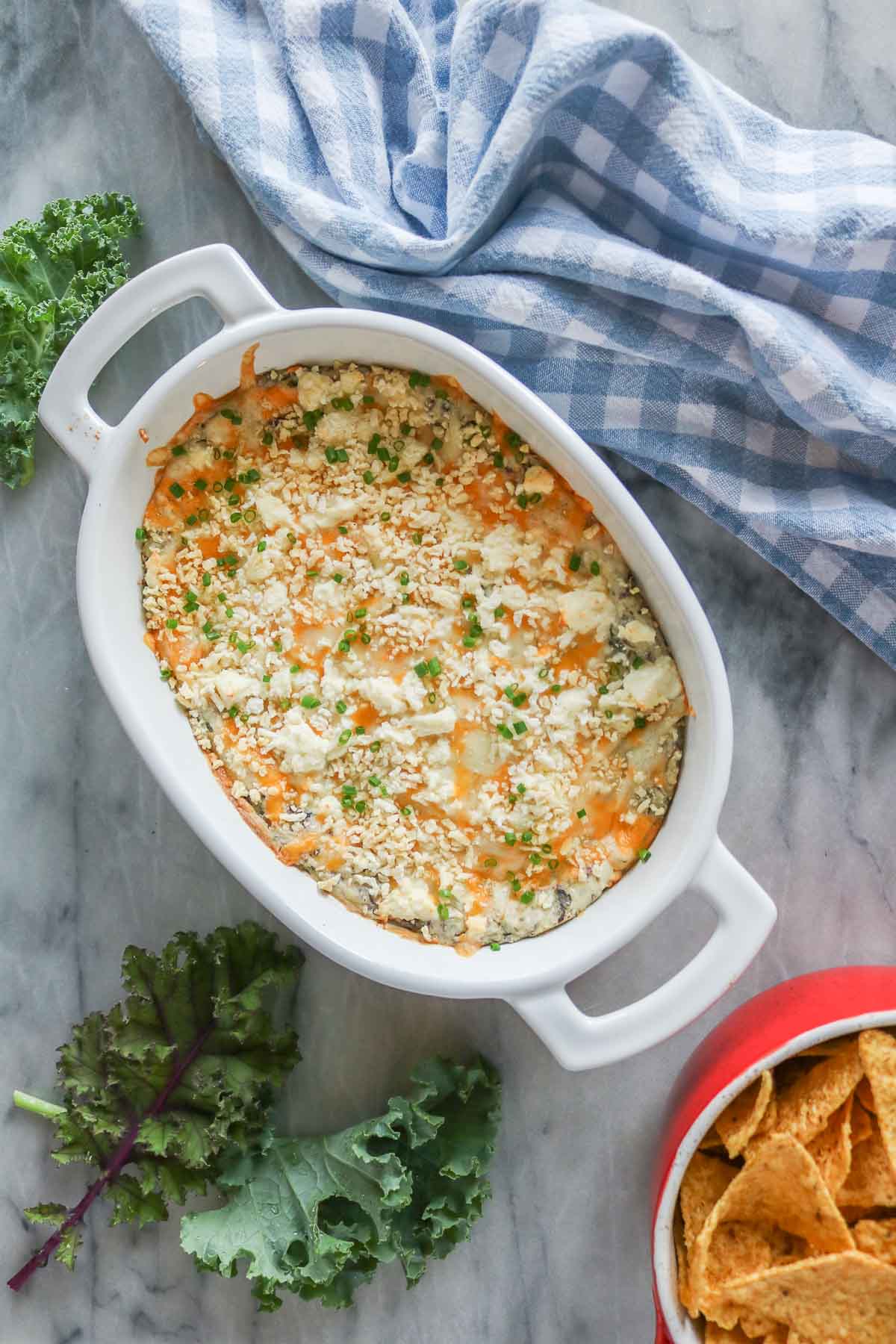 Cheesy kale dip in a baking dish alongside a bowl of tortilla chips and fresh kale.
