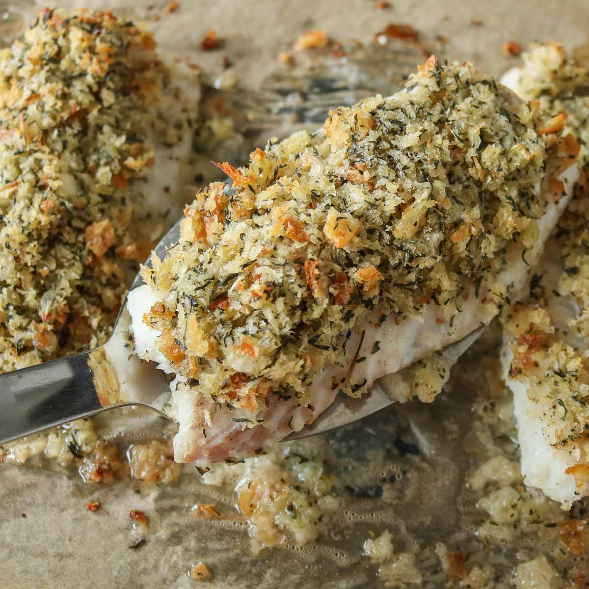 Spatula picking up a baked cod fillet with dressing from a parchment paper-lined sheet pan.