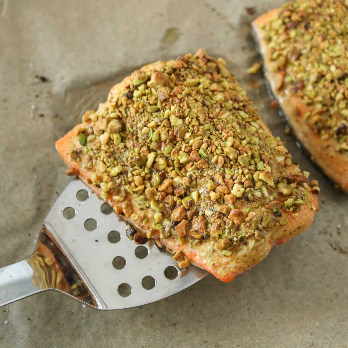 A spatula picking up a baked pistachio-crusted fish fillet from a parchment paper-lined sheet pan.