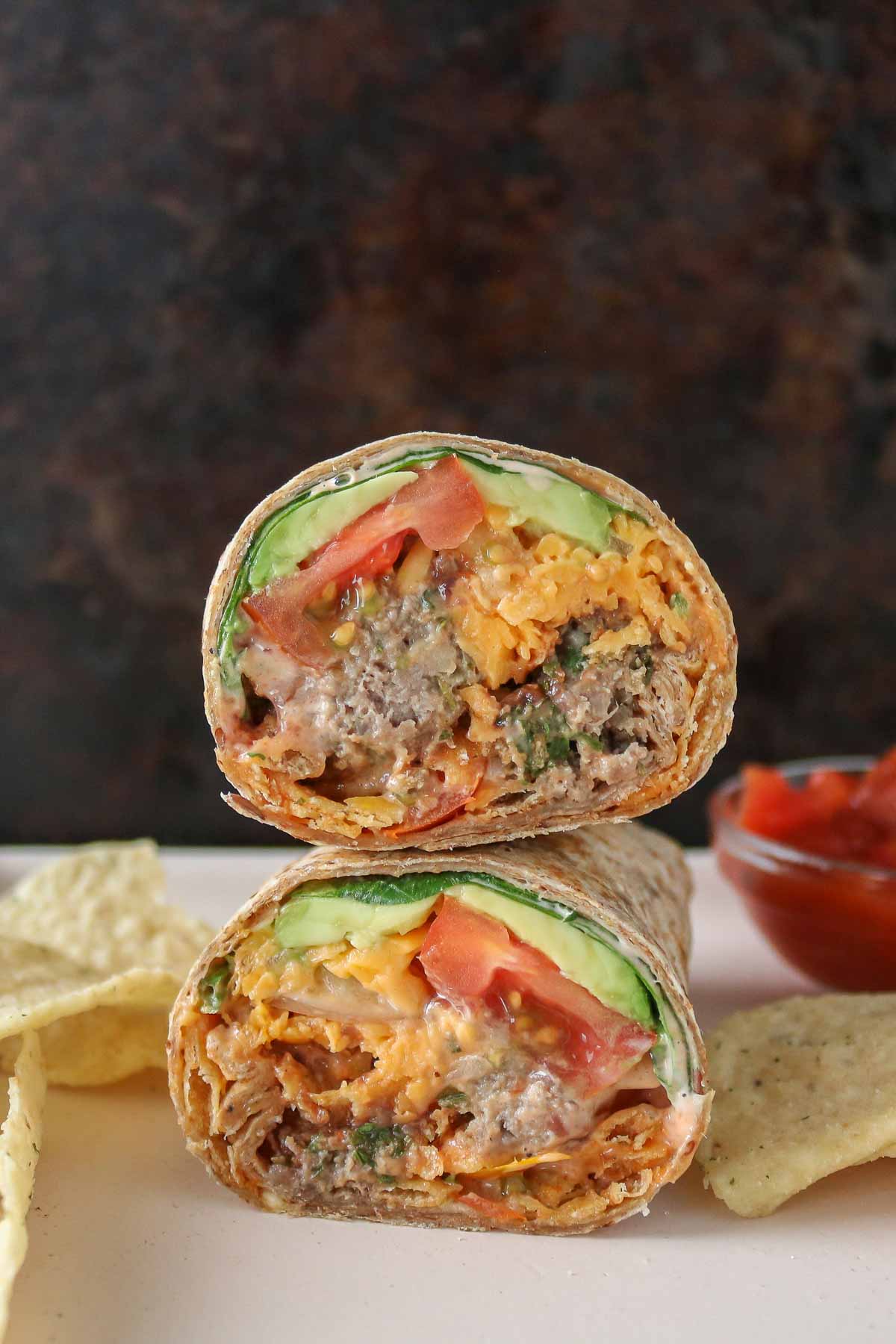 Two halves of a meatball wrap stacked on a plate with tortilla chips and dish of salsa.