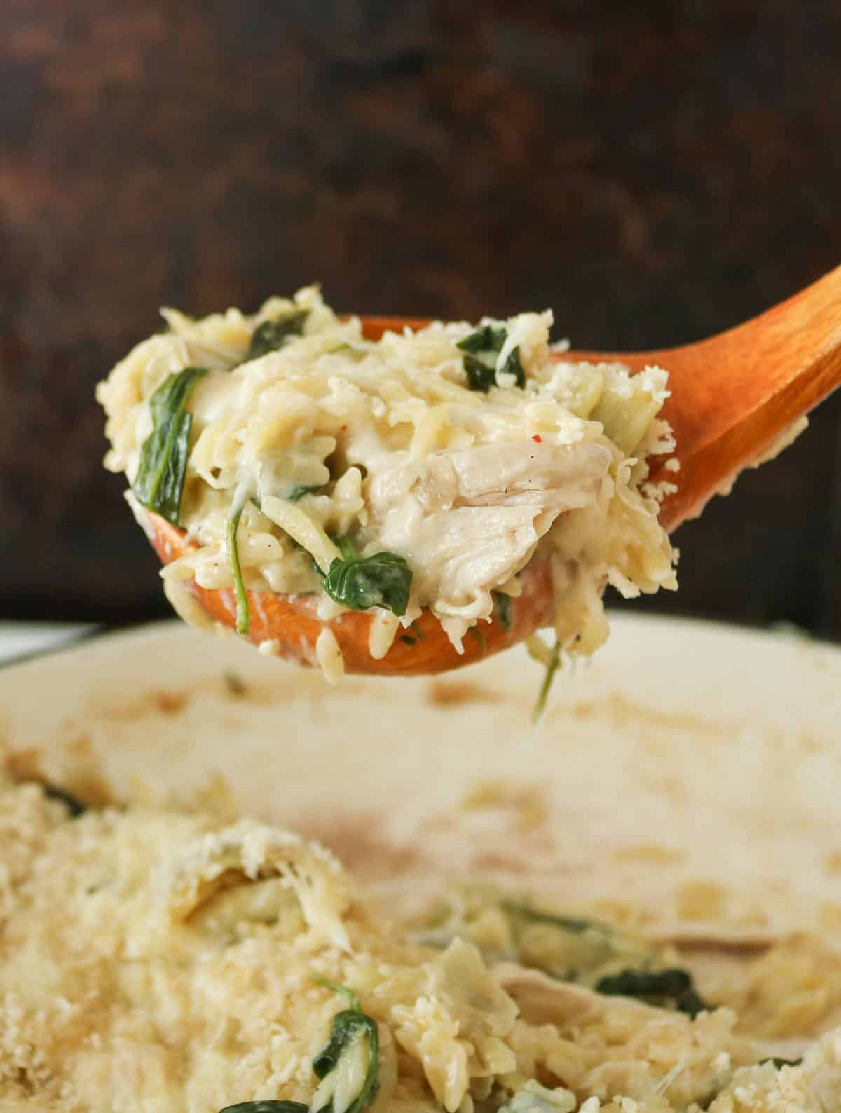 Wooden serving spoon scooping up some orzo bake from a pan.