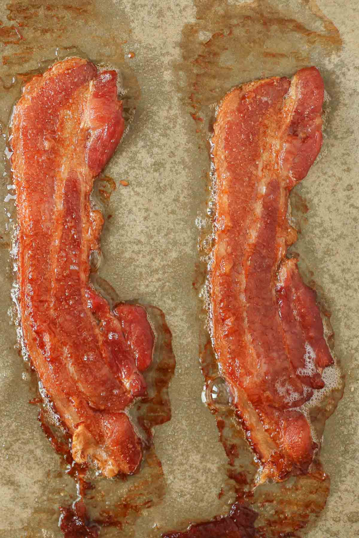 Two slices of crispy thick-cut bacon on a parchment paper-lined sheet pan.