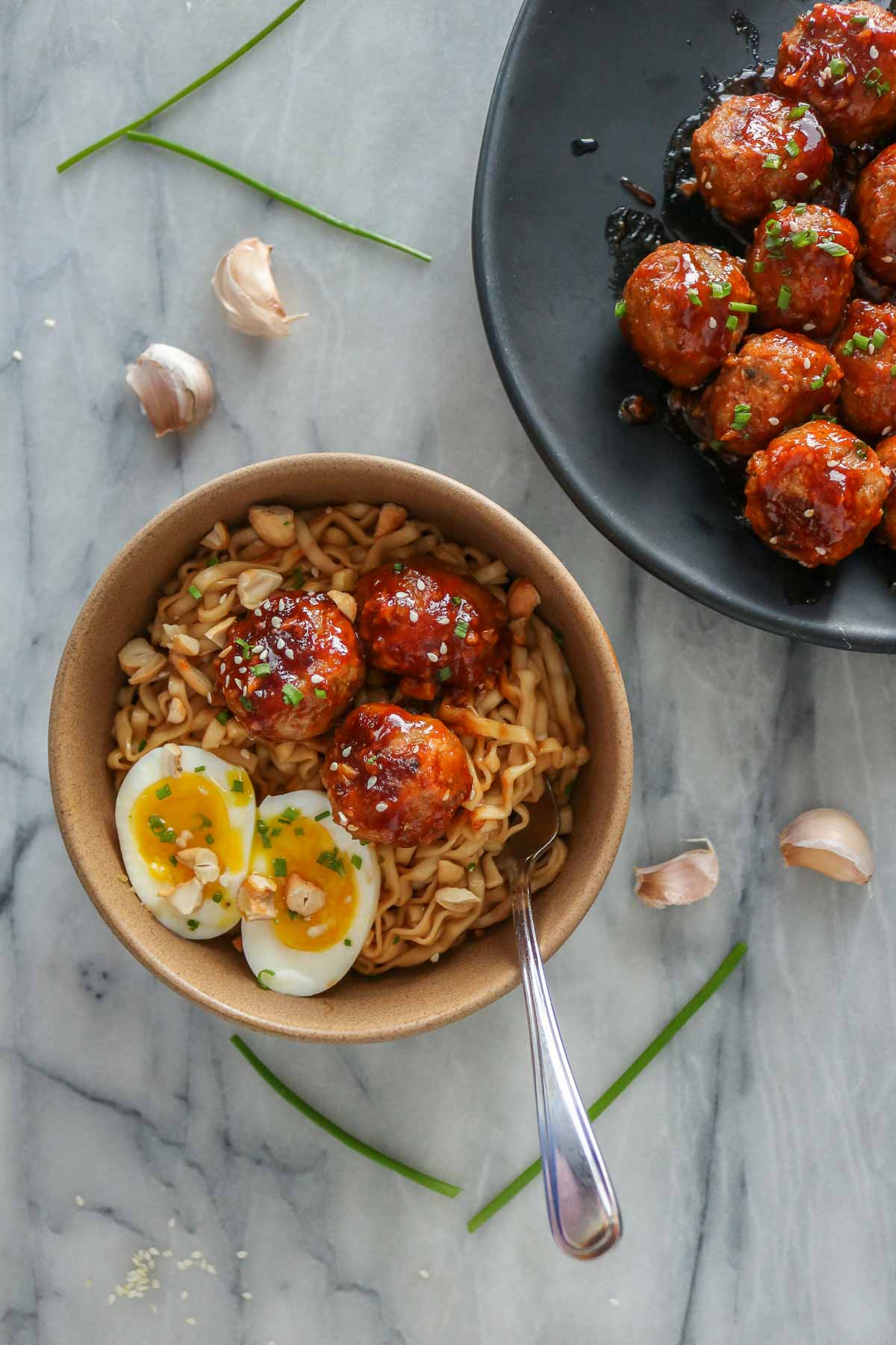 Meatballs, noodles and a jammy soft-boiled egg in a bowl alongside a platter of meatballs.