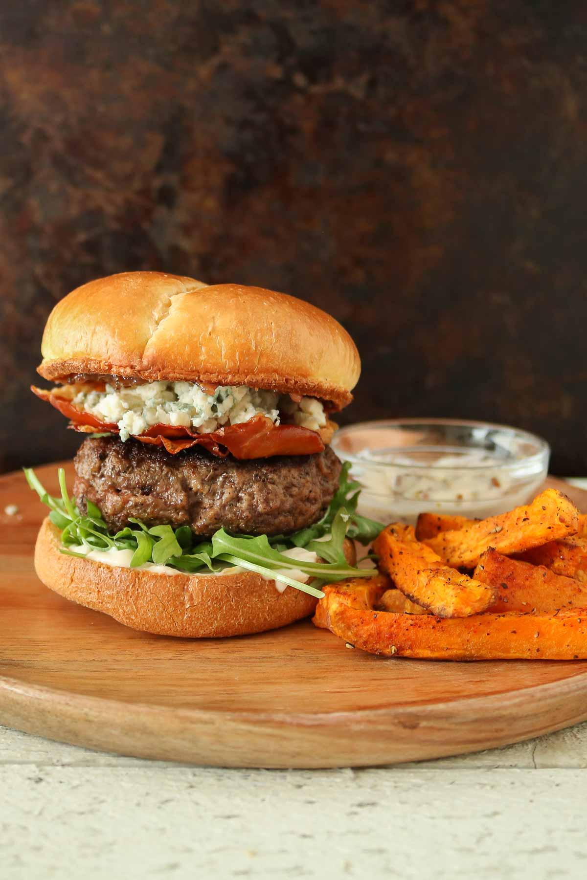 Prosciutto and blue cheese burger, sweet potato fries and dipping sauce on a wooden plate.