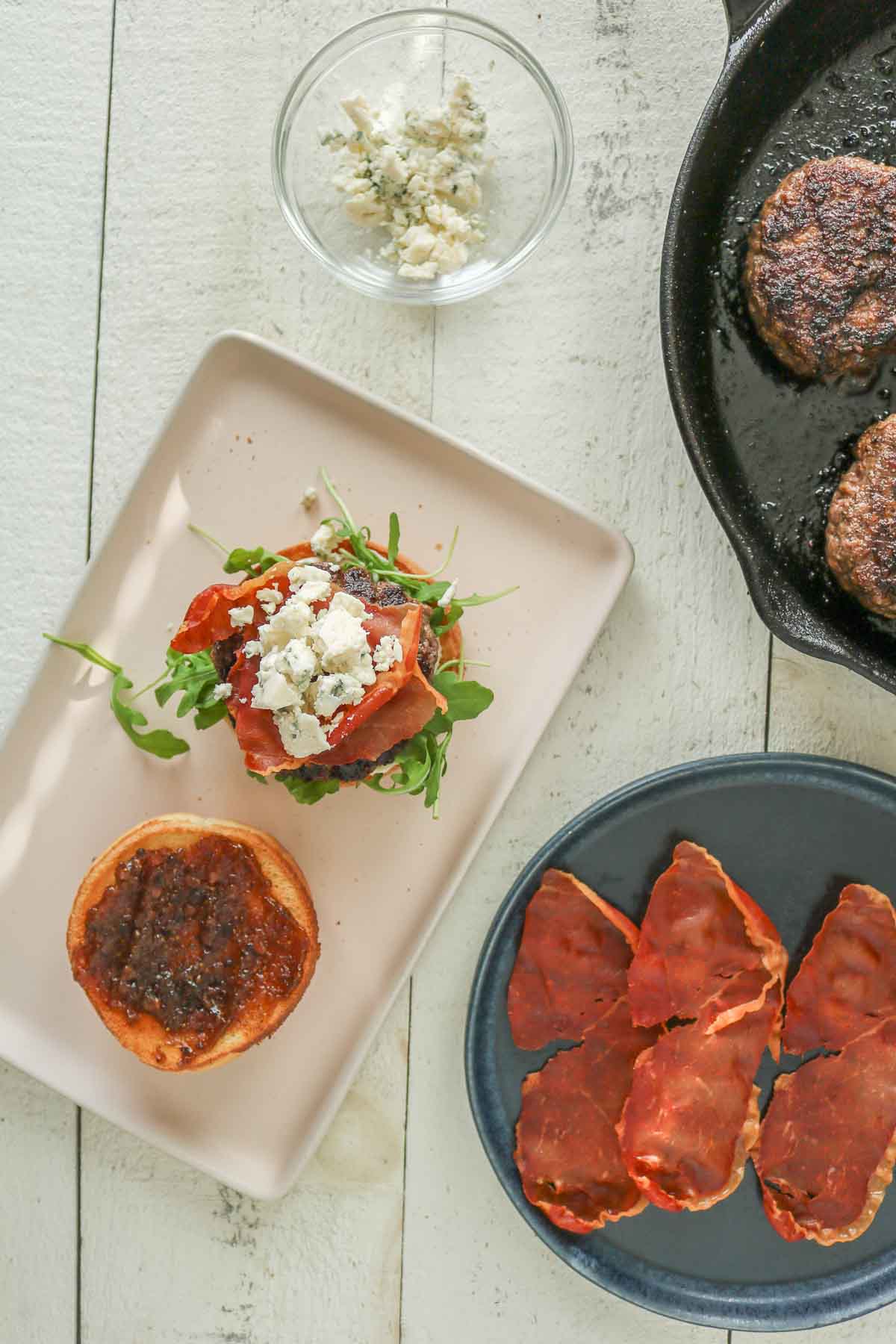 Assembling a homemade burger with crispy prosciutto, blue cheese, arugula and fig jam.