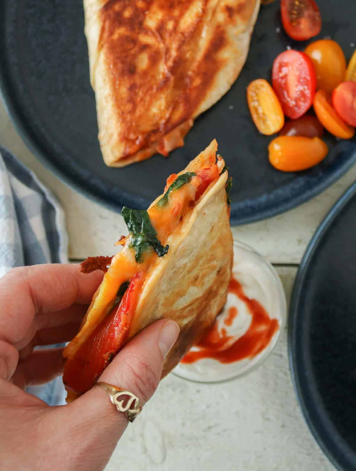 Hand dipping a veggie and egg quesadilla into a small dish of yogurt with hot sauce.