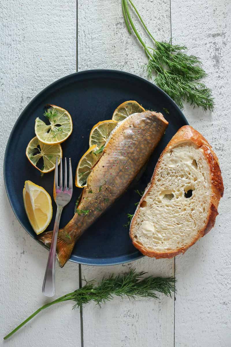 Baked trout with a slice of bread, lemon wedge and a fork on a plate.