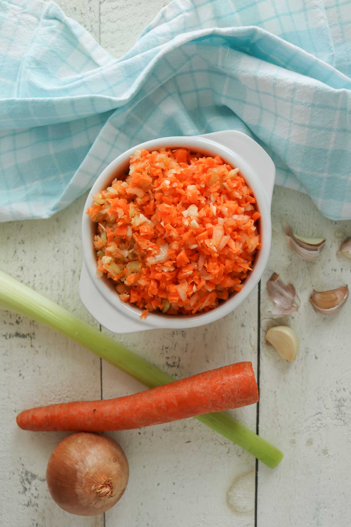 Bowl of finely chopped veggies next to a whole carrot, rib of celery, onion and garlic cloves.