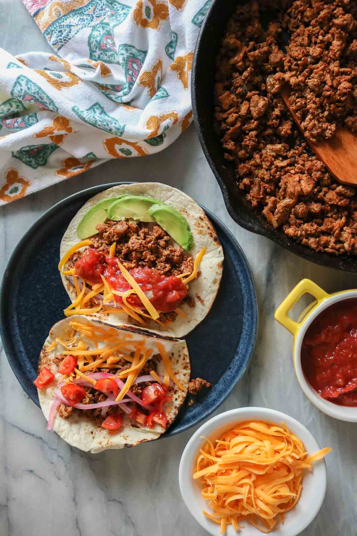 Two chicken tacos on a plate next to a skillet of taco meat and dishes of shredded cheese and salsa.