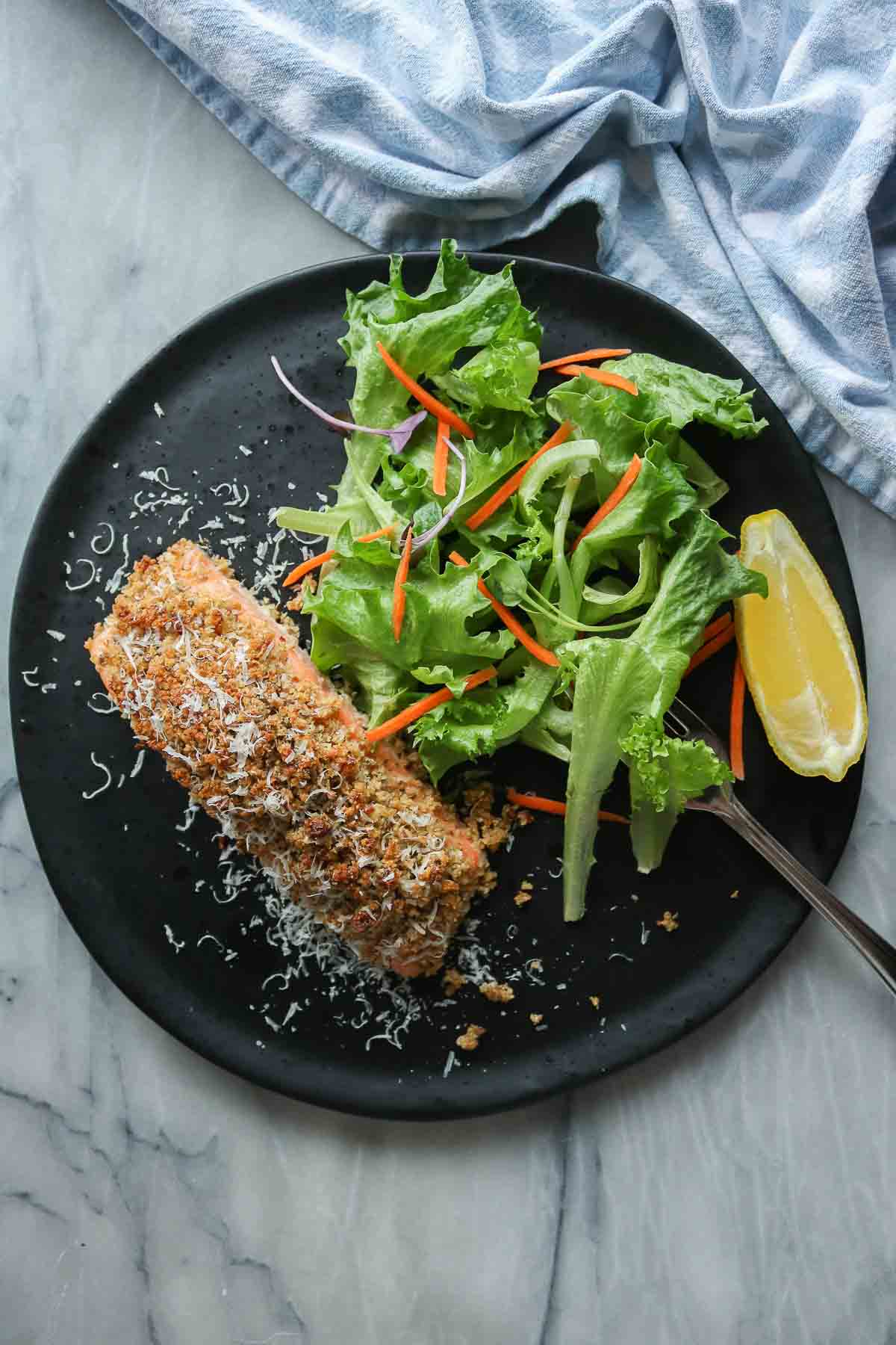 Portion of baked salmon, green salad and lemon wedge on a plate with a fork.