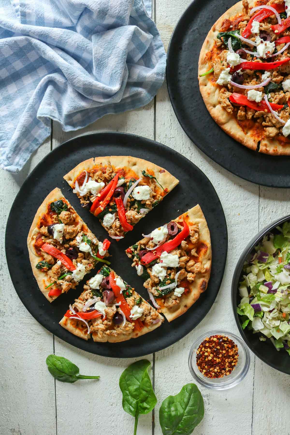 Four slices of pizza on a plate next to a whole pizza, bowl of salad and crushed red pepper flakes.